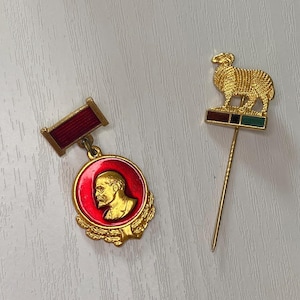 Może przedstawiać: Dwie przypinki w stylu vintage. Jedna to czerwona i złota okrągła przypinka z portretem i czerwonym paskiem. Druga to złota przypinka z owcą na prostokątnej podstawie z brązowymi, czarnymi i zielonymi paskami.