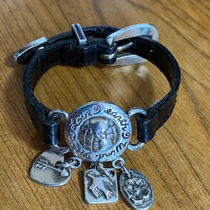 May include: Black leather bracelet with a silver buckle and charms. The charms include a globe with the words "Love Earth World", a heart with an arrow, a dove, and a flower.