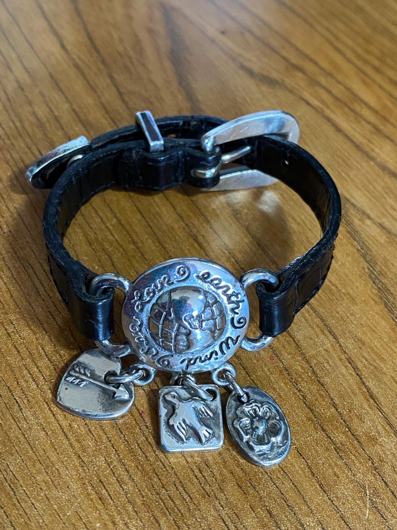 May include: Black leather bracelet with a silver buckle and charms. The charms include a globe with the words "Love Earth World", a heart with an arrow, a dove, and a flower.