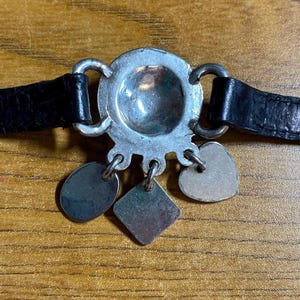 May include: A silver bracelet with a round charm and four dangling charms. The charms are a heart, a square, and two ovals. The bracelet is on a black leather band.