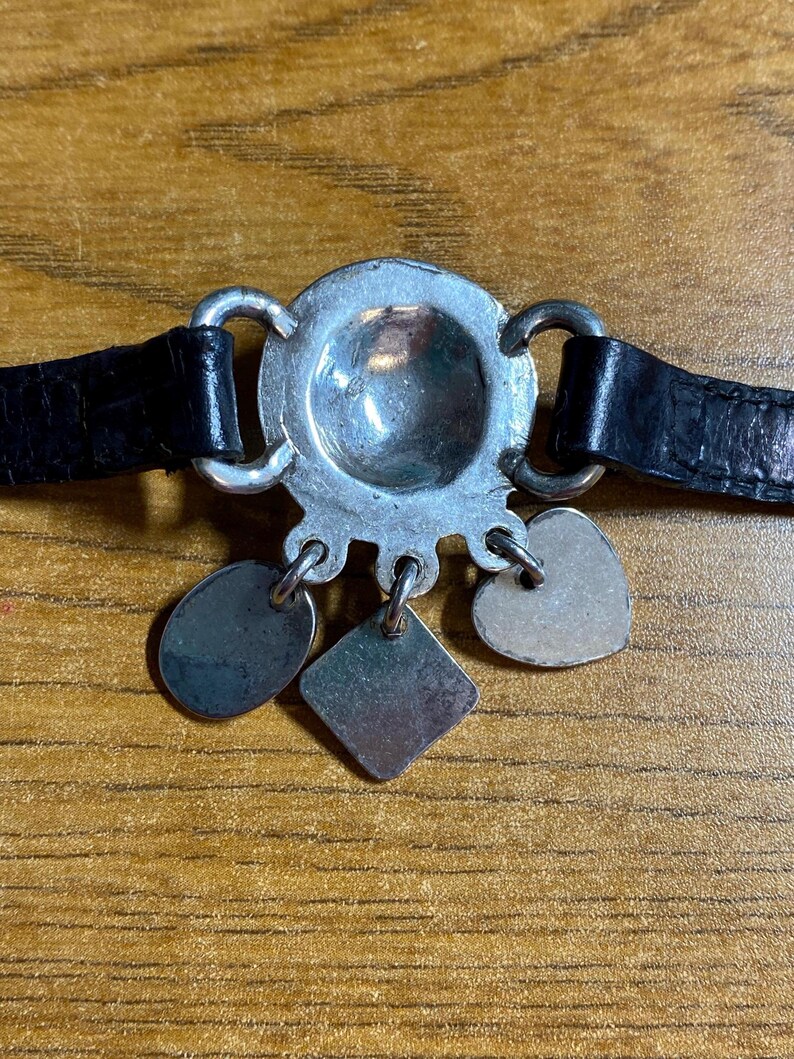 May include: A silver bracelet with a round charm and four dangling charms. The charms are a heart, a square, and two ovals. The bracelet is on a black leather band.