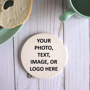 以下が含まれることがあります： 白い丸いコースターに「YOUR PHOTO, TEXT, IMAGE, OR LOGO HERE」という黒い文字が印刷されています。