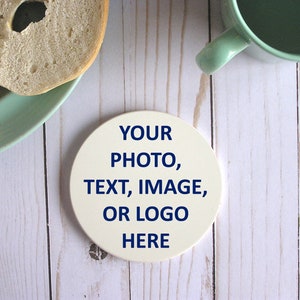 May include: White round ceramic coaster with the text "YOUR PHOTO, TEXT, IMAGE, OR LOGO HERE" in blue lettering.