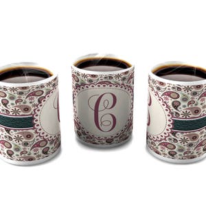 Puede incluir: Tres tazas de café de cerámica blanca con un patrón de paisley en tonos de rosa, verde y marrón. Cada taza tiene una banda azul oscuro alrededor del medio y una gran letra "C" en color burdeos en la parte delantera.