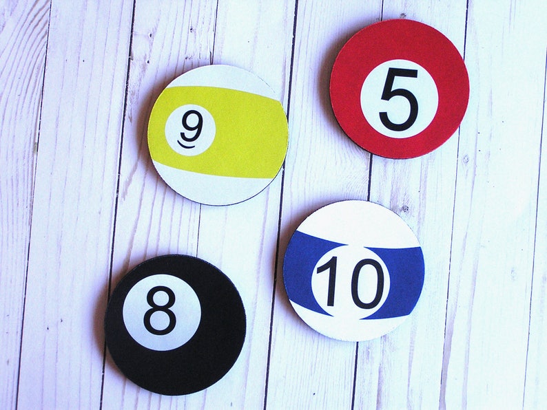 May include: Four round coasters with a white background and black numbers. The coasters are shaped like billiard balls. The numbers on the coasters are 5, 9, 8, and 10. The coasters are on a white wooden surface.