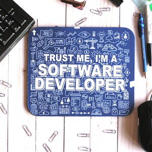 Könnte beinhalten: Ein blaues Mauspad mit weißem Text, der "TRUST ME, I'M A SOFTWARE DEVELOPER" lautet. Das Mauspad ist mit kleinen weißen Symbolen bedeckt, die mit der Softwareentwicklung zusammenhängen.
