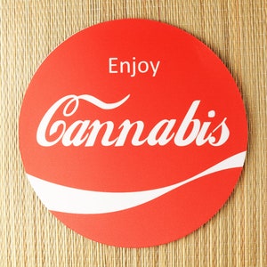 May include: A red circular coaster with the word "Cannabis" in a white, stylized font, with the word "Enjoy" above it. The coaster has a white curved design at the bottom and sits on a woven, tan surface.