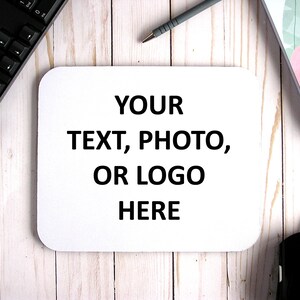 May include: White mouse pad with the text "YOUR TEXT, PHOTO, OR LOGO HERE" printed in black on a wooden surface.