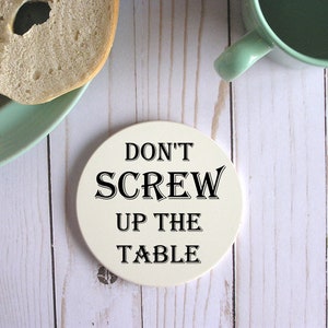 Op de afbeelding: Ronde witte onderzetter met zwarte tekst die "Don't Screw Up The Table" zegt.