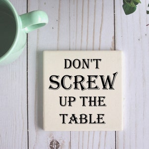 Op de afbeelding: Witte keramische onderzetter met zwarte tekst die "Don't Screw Up The Table" zegt.