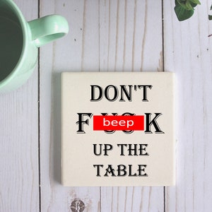Op de afbeelding: Witte keramische onderzetter met zwarte tekst die "DON'T [beep] UP THE TABLE" luidt.
