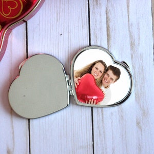 May include: A silver heart-shaped compact mirror with a hinged lid. The mirror is open, revealing a photo of a couple holding a red heart-shaped balloon. The couple is smiling and looking at the camera.