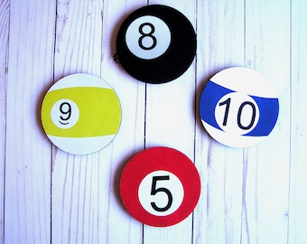 May include: Four round pool ball coasters with white backgrounds and black numbers. The coasters are 5, 8, 9, and 10. The coasters are on a white wood background.