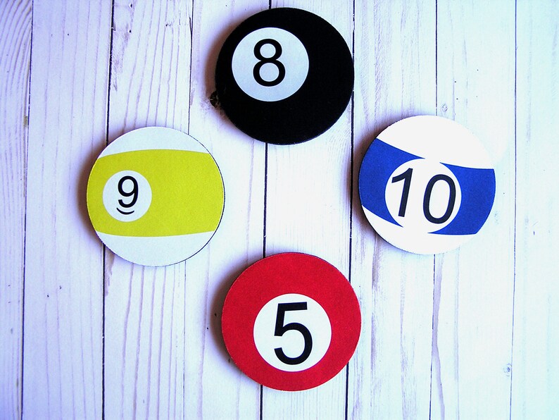May include: Four round pool ball coasters with white backgrounds and black numbers. The coasters are 5, 8, 9, and 10. The coasters are on a white wood background.