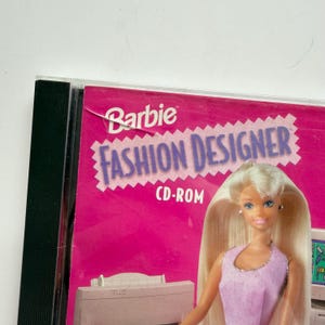 May include: A Barbie Fashion Designer CD-ROM case featuring a bright pink cover with the text Barbie Fashion Designer CD-ROM. The cover displays a graphic of a blonde doll wearing a light purple sleeveless top, positioned above a computer printer.