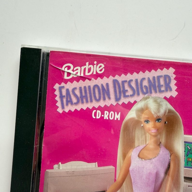 May include: A Barbie Fashion Designer CD-ROM case featuring a bright pink cover with the text Barbie Fashion Designer CD-ROM. The cover displays a graphic of a blonde doll wearing a light purple sleeveless top, positioned above a computer printer.