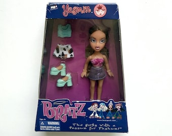 May include: A Bratz Yasmin doll in original packaging, featuring a doll with long brown hair wearing a pink fuzzy top and denim skirt. Included accessories are a light blue tank top, a patterned skirt, and two light blue platform shoes.