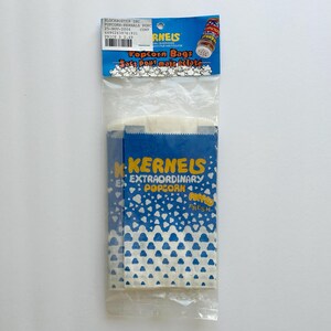May include: A package of three white popcorn bags with blue and white hearts printed on them. The bags are labeled "KERNELS EXTRAORDINARY POPCORN" and "POPPED FRESH".
