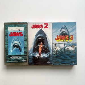 Peut inclure: Trois cassettes VHS du film Jaws disposées côte à côte. La première montre un requin remontant vers un nageur avec le titre Jaws. La deuxième montre un requin derrière un skieur nautique avec le titre Jaws 2. La troisième montre une tête de requin.