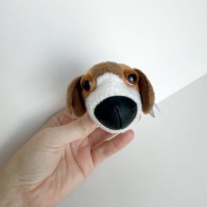 May include: A plush dog head toy featuring brown and white fur, large dark brown eyes, and a prominent black plastic nose. The soft toy has floppy ears and a white patch extending from the forehead down to the muzzle, creating a realistic canine appearance.
