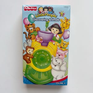 May include: A Fisher-Price "Little People Discovering Animals" VHS tape. The cover shows cartoon animals like a tiger, giraffe, elephant, and pig, with the title and "Volume 3" text. The background is light blue.