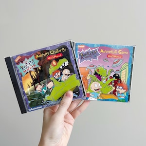 May include: Two Rugrats video game instruction manuals. The left manual is purple, blue, and green with the text "The Rugrats Movie Activity Challenge User's Manual" and a picture of the Rugrats characters riding a green monster. The right manual is purple, blue, and green with the text "Rugrats Adventure Game User's Manual" and a picture of the Rugrats characters with a green dinosaur.