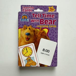 May include: A purple box of Bear in the Big Blue House Tell Time With Bear learning cards for ages 2-5. The box features a brown bear character and two sample cards showing a clock face at 8:00 and the text 8:00 eight o'clock.