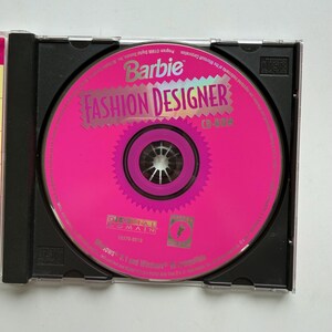May include: A Barbie Fashion Designer CD-ROM disc housed in a black plastic jewel case. The circular disc is bright pink with a jagged-edged center design, featuring the text Barbie Fashion Designer, Digital Domain, and Windows 3.1 and 95 compatible.