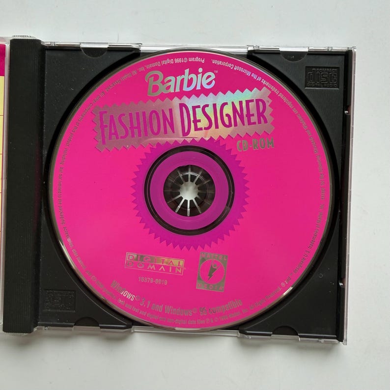 May include: A Barbie Fashion Designer CD-ROM disc housed in a black plastic jewel case. The circular disc is bright pink with a jagged-edged center design, featuring the text Barbie Fashion Designer, Digital Domain, and Windows 3.1 and 95 compatible.