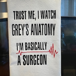 May include: White kitchen towel with the text "TRUST ME, I WATCH GREY'S ANATOMY I'M BASICALLY A SURGEON" in black lettering. A red EKG line is printed next to the text. The towel is hanging on a metal bar.