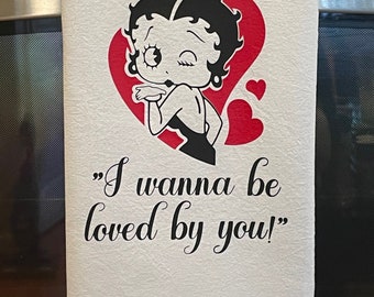 Op de afbeelding: Witte keukendoek met een cartoonafbeelding van Betty Boop die een kus blaast, tegen een rode hart. De handdoek heeft de zin "I wanna be loved by you!" gedrukt in zwart schrift.