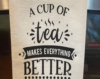 May include: White kitchen towel with the text "A CUP OF tea MAKES EVERYTHING BETTER" in black. The towel is hanging on a stainless steel appliance. The text is in a decorative font with steam and star accents.