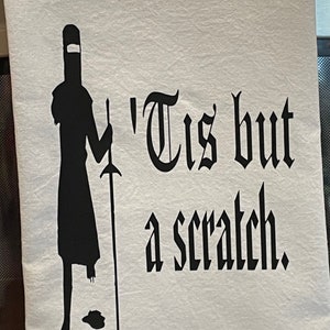 May include: White kitchen towel with a black silhouette of a knight holding a sword, with the text "'Tis but a scratch." The knight is missing a leg. The towel is likely made of cotton.