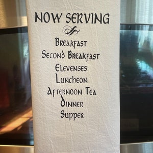 May include: White kitchen towel with black text that reads "NOW SERVING" followed by a list of meal times: Breakfast, Second Breakfast, Elevenses, Luncheon, Afternoon Tea, Dinner, and Supper. The towel is hanging on a stainless steel appliance.