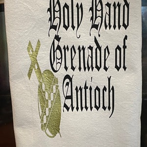 May include: White kitchen towel with the phrase "Holy Hand Grenade of Antioch" in black gothic lettering. A gold-colored illustration of a grenade and cross is on the left side of the text.
