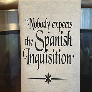 May include: White kitchen towel with the phrase "Nobody expects the Spanish Inquisition" in black script. A black floral design is below the text. The towel is hanging on a stainless steel appliance.