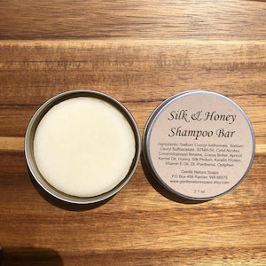 Può includere: Una saponetta per capelli rotonda e bianca in una scatola di latta argentata con un'etichetta marrone che recita "Silk & Honey Shampoo Bar" e elenca gli ingredienti. L'etichetta include anche il nome dell'azienda, l'indirizzo, il sito web e la dimensione della saponetta, che è di 2,1 once.