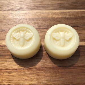 Puede incluir: Dos barras de jabón redondas, blancas y sólidas con un diseño de abeja en relieve en la parte superior. La abeja está rodeada de un círculo.