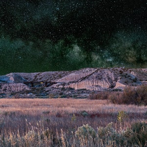 May include: A landscape photograph of a grassy field with a hill in the background. The sky is dark and filled with stars, including the Milky Way. A full moon is visible in the upper right corner of the image.