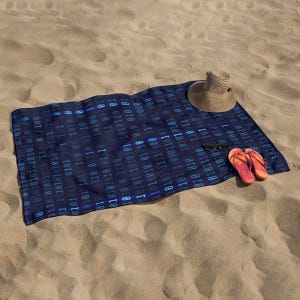 May include: A navy blue beach towel with a binary code pattern, a straw hat, sunglasses, and orange flip-flops on a sandy beach. The towel is rectangular and has a repeating pattern of blue binary digits.