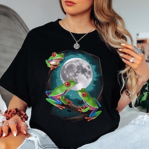 May include: Black t-shirt featuring a vibrant graphic of three green tree frogs with red eyes, perched on a branch in front of a full moon. The design has a whimsical, nature-inspired theme.