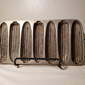 May include: Vintage cast iron cornbread pan with six individual molds shaped like ears of corn. The pan is silver in color and has a black metal stand.