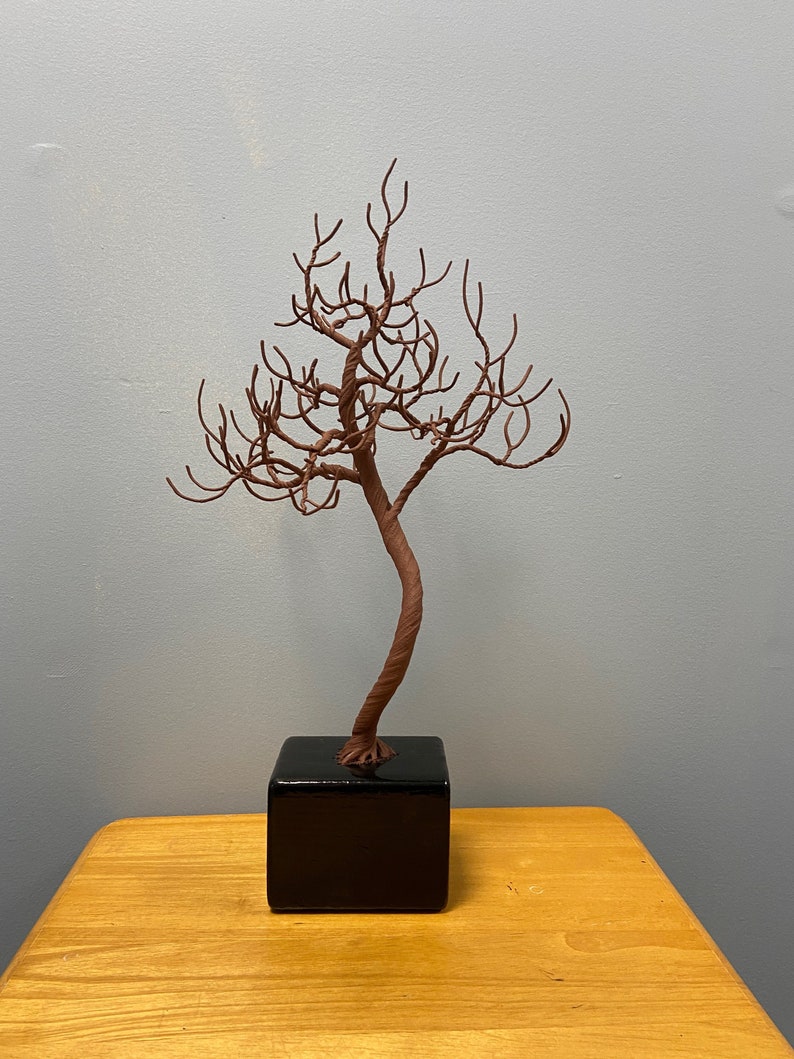 Puede incluir: Una escultura de alambre de un &aacute;rbol con un acabado marr&oacute;n, de pie sobre una base cuadrada negra. El &aacute;rbol est&aacute; hecho de finas ramas de alambre que est&aacute;n retorcidas y giradas para crear un aspecto realista.