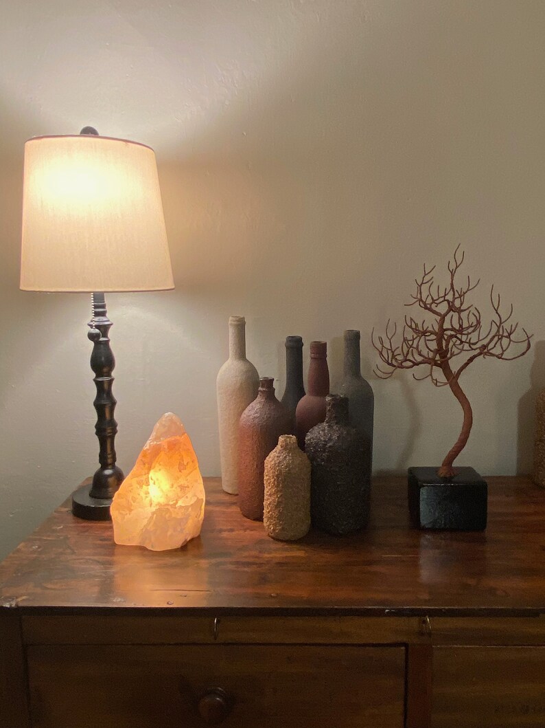 Puede incluir: Una mesa de madera con una l&aacute;mpara, una l&aacute;mpara de sal y una colecci&oacute;n de botellas decorativas. Un peque&ntilde;o &aacute;rbol de ramas secas se encuentra sobre la mesa.