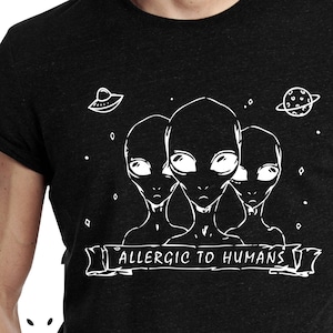 Puede incluir: Camiseta gris oscuro con un gráfico blanco de tres caras alienígenas, una nave espacial y un planeta. El texto "ALLERGIC TO HUMANS" se muestra en una pancarta. La camiseta es de cuello redondo.