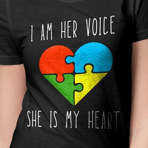 Puede incluir: Camiseta negra con texto blanco que dice "Soy su voz" y "Ella es mi corazón". Un corazón de pieza de rompecabezas colorido se encuentra en el centro del texto.