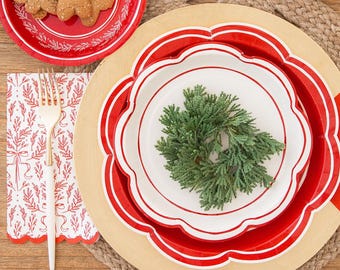 May include: A festive holiday table setting with red and white plates, gingerbread cookies, and a small house-shaped gift box. The table is decorated with evergreen branches, gold cutlery, and a glass of liquid.