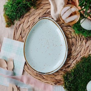 May include: A light blue speckled paper plate with a gold rim sits on a woven straw placemat surrounded by moss and pastel blue and white checked napkins.