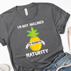 May include: A dark gray t-shirt with the text "I'm not inclined to resign to maturity" and a yellow pineapple graphic with a white banner that says "to resign to".
