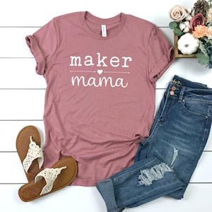 May include: A dusty rose t-shirt with the text "maker mama" in white with a small heart between the words. The shirt is laying on a white surface with a pair of tan sandals, blue jeans, and a small white pumpkin in a wooden box with pink flowers.